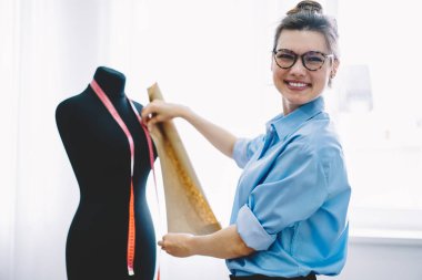 Neşeli kadın terzi kameraya bakıyor. Elinde mezurayla mankenin yanında duruyor. Atölyede kıyafet dikme sırasını yerine getirirken ölçümler yapıyor.