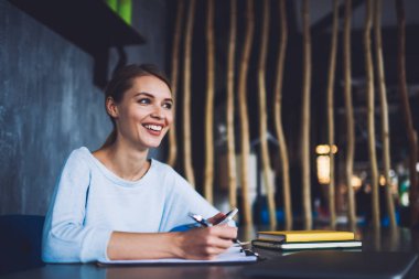 Genç, gülümseyen bir bayan, ofiste çalışırken elinde cep telefonuyla, elinde not defterleriyle masada oturuyor ve mutlu mesut uzaklara bakıyor.