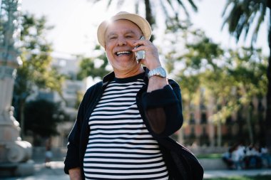 Hasır şapkalı neşeli erkek turistin yarım boy portresi modern akıllı telefon cihazıyla olumlu uluslararası iletişim sırasında gülüyor.