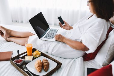 Beyaz sabahlık giymiş, beyaz elbiseli, akıllı telefonuyla mesajlaşan ve Kuala Lumpur 'da dizüstü bilgisayarla yatakta dinlenen bir kadının yan görüntüsü.