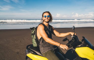 Cheerful bearded male motorcyclist in undershirt sitting on motorbike among tropical beach and looking at camera while enjoying summer holidays