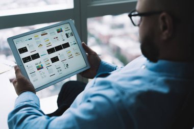 Resmi gömlek ve gözlüklü tanınmayan girişimcinin arka planı karanlık ofis odasındaki pencerenin yanında tablet kullanırken ve bilgileri kontrol ederken