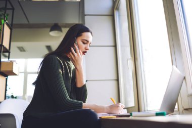 Pencerenin kenarında dizüstü bilgisayarla oturan ve iş yerindeki cep telefonuyla konuşurken notlar yazan kendini ifade eden kadın girişimcinin düşük açısı