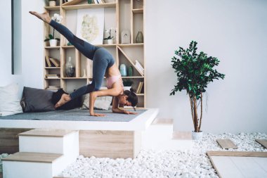 Spor kıyafetli konsantre genç bir kadının podyumda yoga yaparken tek bacaklı bir karga pozisyonunda duruşu.