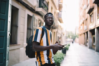 Akıllı telefon cihazıyla düşünceli hippi adam şehir sokaklarını düşünürken dikkatle başka yere bakıyor.