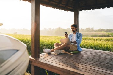 Çapraz bacaklı, günlük kıyafetleriyle neşeli adam ahşap terasta oturmuş akıllı telefondan internette sörf yapıyor ve bilgisayarını karıştırıyor.