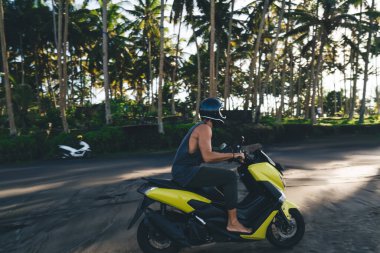 Kimliği belirsiz erkek motosikletçinin koruyucu kaskı ve günlük kıyafetleri var. Motosiklet sürüyorlar. Palmiye ağaçlarının yakınındaki kumsalda.
