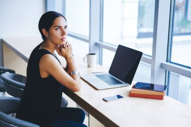 Uzaktan kumandalı sakin kadın işçinin yan görüntüsü. Koyu saçlı, başka tarafa bakıyor ve iş projesini düşünüyor. Çağdaş çalışma alanında masada otururken.