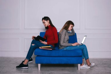 Beyaz salondaki mavi kanepede sırt sırta otururken günlük kıyafetlerini giymiş ev ödevi yapan tüm kadınların yan görüntüsü.