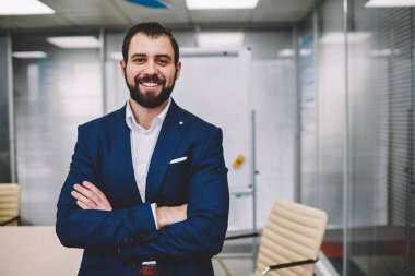 Çapraz kolları ve dişlek gülümsemesiyle modern çalışma alanında çalışan ve kameraya bakan kendine güveni tam bir erkek yönetici.