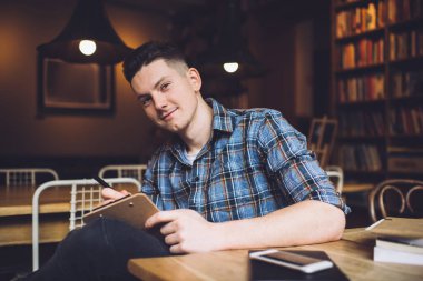 Elinde kalem ve panoyla gülümseyen bir erkek rahat kütüphanede elinde kitaplarla oturmuş kameraya bakıyor.