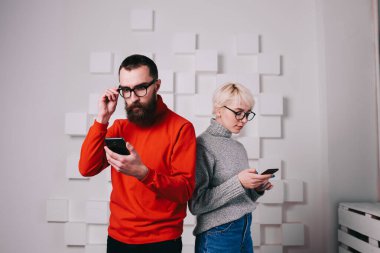 Turuncu süveter ve gözlüklü genç sakallı hippi cep telefonundan mesaj okuyor ve düşünceli genç bir bayanın yanında durup akıllı telefonu karıştırırken şaşırmış görünüyor.