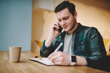 Gülümseyen genç bir erkek iş arayan cep telefonuyla konuşuyor ve bir bardak sıcak içecekle masaya oturmuş not alıyor.