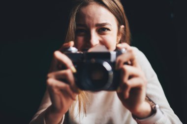 Beyaz kazaklı, genç, neşeli kadın fotoğrafçı. Eski moda dijital fotoğraf makinesiyle fotoğraf çekiyor. Karanlık odada kameraya bakıyor.