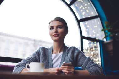 Kafeteryada bir fincan kahve ve cep telefonuyla oturan düşük açılı, odaklanmış genç bir kadın.