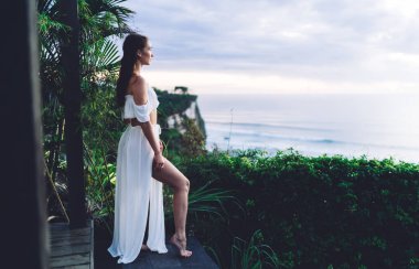 Çıplak ayaklı, şık giyinmiş, egzotik bitkilere dalmış, terasın önünde dikilmiş ve gözlerini kaçıran genç bir kadın.