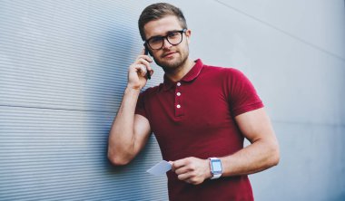 Günlük kıyafetleri ve kol saati takan sakallı sakin bir adam kartvizitini tutuyor ve gün ışığında gri duvarda cep telefonuyla konuşuyor.