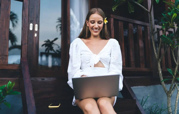 Hafif giyimli memnun iş kadını basamaklarda dizüstü bilgisayarla oturuyor ve tropikal bir ülkede dinlenirken yeni bir proje üzerinde çalışıyor.