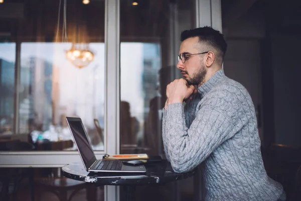 Side view of serious bearded male employee browsing netbook while sitting at table in cozy cafe and working remotely on project