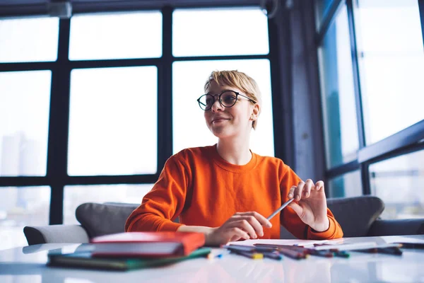 From below of happy female in casual clothes with eyeglasses looking away sitting at table near window with colored pencils while working remotely
