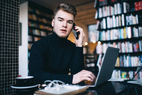 Düşünceli genç adam telefonda konuşuyor ve kütüphanede dizüstü bilgisayarın önünde otururken kameraya bakıyor.