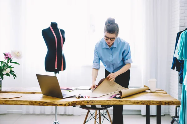 現代的なアトリエで仕事中の新しい服のスケッチとクラフト紙を見てテーブルの近くに立つ若い焦点を当てた女性のシームレスストレス — ストック写真