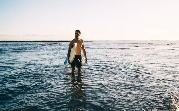 身穿泳衣 身披长衫 身披紧身衣的年轻男子运动员 在日落时分 独自站在海浪中 手握冲浪板 望向远方 — 图库照片