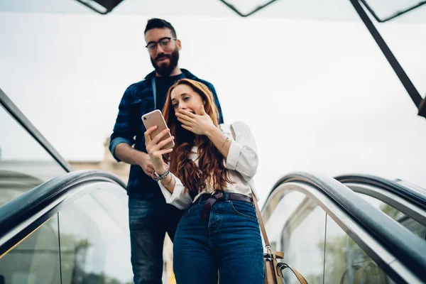 Amazed hipster girl reading text publication with shocked news sharing content with blurred boyfriend