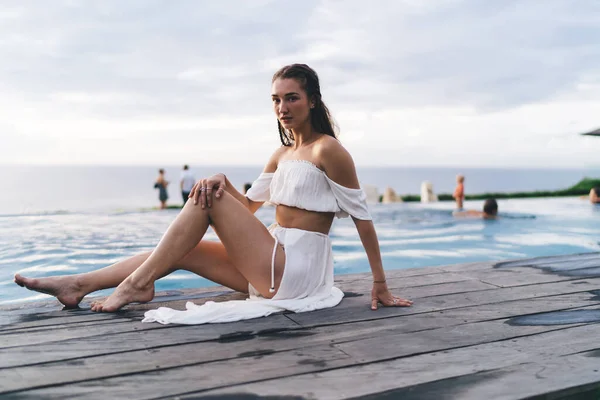 Uzun boylu, çıplak ayaklı, düşünceli, zarif elbiseli bir kadın yazın tahta havuz başında otururken kameraya bakıyor.