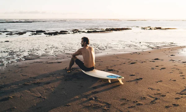 阳光明媚的夜晚 穿着短裤坐在冲浪板上 欣赏沙滩上平静大海之美的裸男冲浪运动员的背影 — 图库照片
