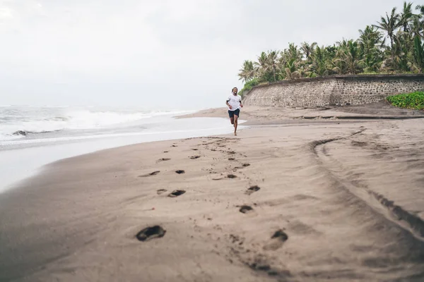Full body African American male jogger in sportswear running on sandy beach near raging sea waves during cloudy summer day at tropical resort