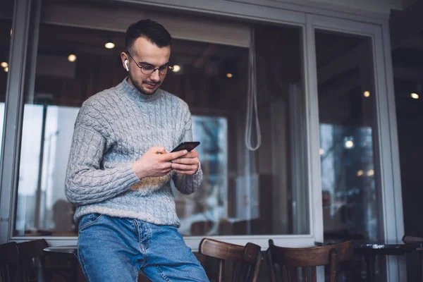 Kablosuz kulaklık ve gözlüklü genç, olumlu sakallı adam. Pencere kenarında otururken ve dinlenirken cep telefonu kullanıyor.