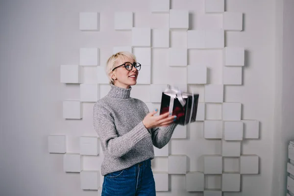 Delighted woman in turtleneck and eyeglasses smiling and holding in hands gift box while spending time in white modern apartment