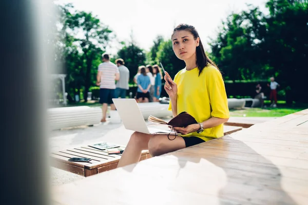 Young woman in casual clothes sitting on wooden bench with laptop and planner and looking thoughtful while working on project remotely