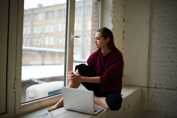 穿着休闲装的认真的女自由职业者一边坐在窗台上一边看着一边 一边舒服地拿着笔记本电脑 梦游般地看着远方 — 图库照片