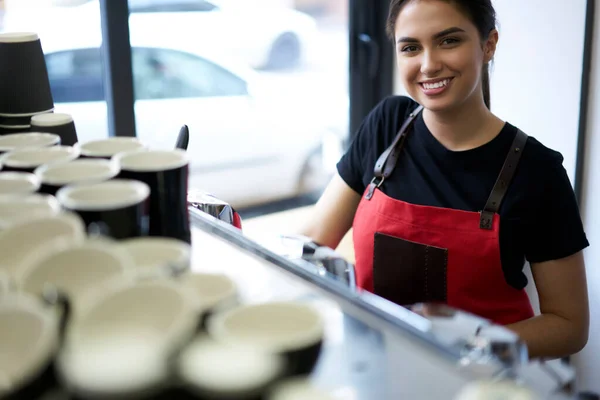 Half length portrait of cheerful caucasian woman in apron looking at camera working at coffee shop using equipment