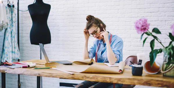 Yeni projede çalışırken müşteriyle konuşan genç kadın terzi ahşap masada çizimler ve kalemlerle oturuyor.