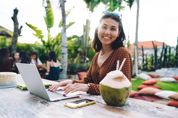Cheerful Asian female in casual outfit sitting at wooden table with coconut drink and typing on laptop keyboard in cafe
