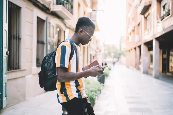 Side view of millennial dark skinned tourist with travel backpack using cellular application for tracking location GPS