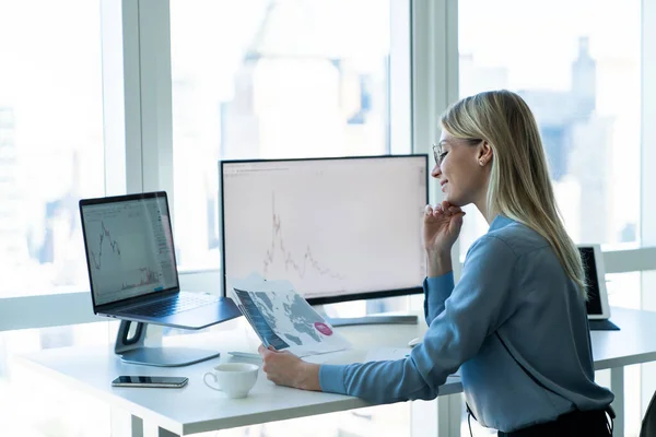 Side view of positive young female employee in formal clothes and eyeglasses sitting at desk in light office and checking documents while working
