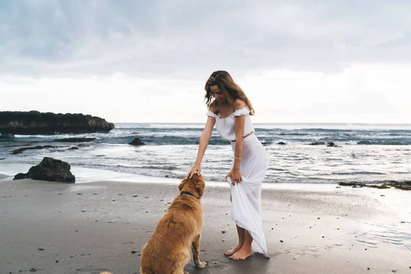 Full body delighted barefoot female in casual wear standing on wet sandy seashore and caressing obedient dog while enjoying stroll