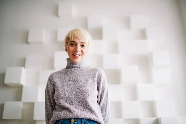 Low angle of young cheerful female in casual sweater with toothy smile and hands behind back looking at camera standing near wall with cube