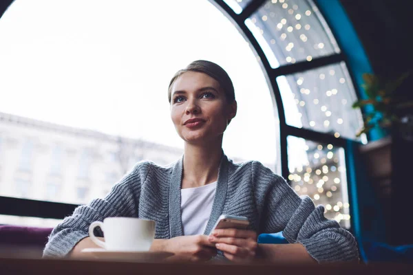 Kafeteryada bir fincan kahve ve cep telefonuyla oturan düşük açılı, odaklanmış genç bir kadın.