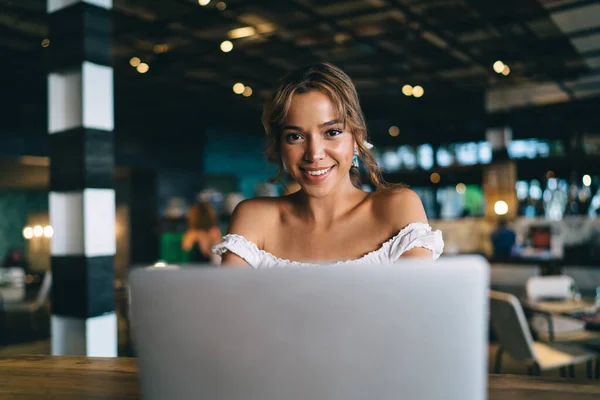 Smiling young lady sitting at table in cafe and using laptop for remote work and wearing casual clothes while looking at camera
