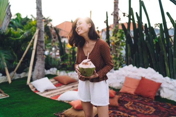 Young ethnic female in summer blouse and shorts smiling happily with coconut water in hands while standing on open terrace of resort house