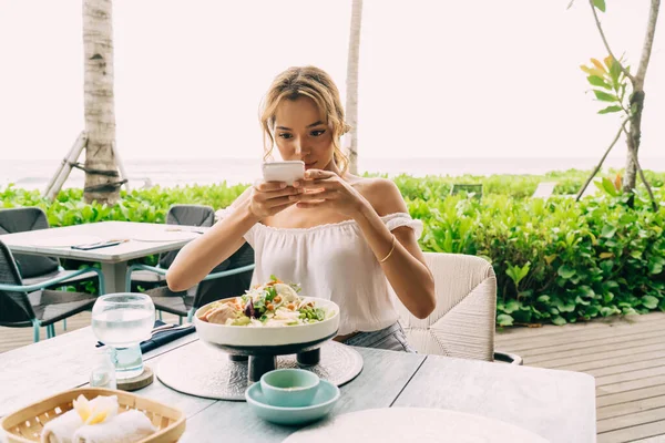 Sakin bir kadın masada oturmuş, açık kafede yemek yerken akıllı telefondan lezzetli yemeklerin fotoğrafını çekiyor.