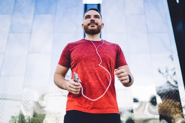 From below of young bearded man in sportswear and earphones holding mobile phone while jogging on street in summer day