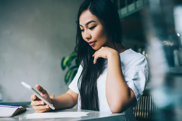 年轻的亚洲女学生穿着休闲装 坐在餐桌前 一边用智能手机做作业 一边在现代咖啡馆里做作业 — 图库照片