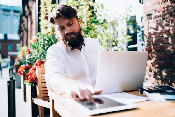 Kafe 'de açık havada çalışırken netbook ile masada çalışırken tablet üzerinde interneti tarayan odaklanmış erkek uzaktan kumanda işçisi.