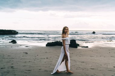 Beyaz elbiseli genç etnik kadın gezginin tüm vücudu dalgalı deniz kenarındaki kumlu sahil boyunca yürüyüp kameraya bakarken.